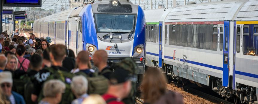 Tłum ludzi na dworcu, pociąg wjeżdżający na peron. Słoneczny dzień, niebo bez chmur.