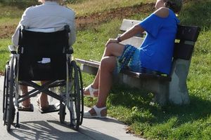 Dwie kobiety siedzą na ławce, jedna w wózku inwalidzkim, obok zielona trawa i ścieżka.
