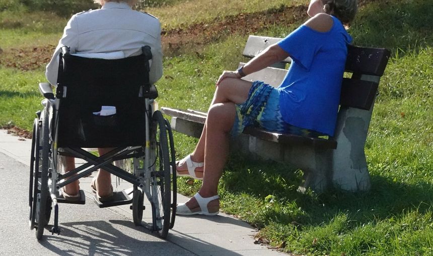 Dwie kobiety siedzą na ławce, jedna w wózku inwalidzkim, obok zielona trawa i ścieżka.