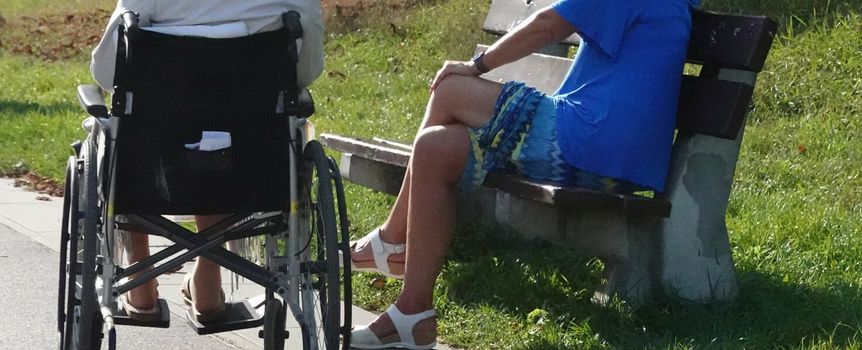 Dwie kobiety siedzą na ławce, jedna w wózku inwalidzkim, obok zielona trawa i ścieżka.