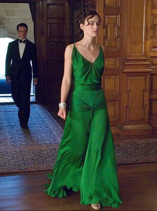 Młoda kobieta w zielonej sukni idzie w eleganckim wnętrzu, za nią mężczyzna w garniturze.