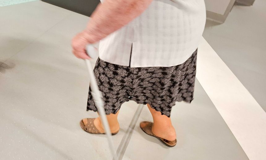 Kobieta w białej bluzce i czarnej spódnicy, idąca z laską po gładkiej podłodze.