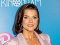 Joanna Jabłczyńska z krótkimi włosami, uśmiechnięta, w różowej marynarce, na jasnoniebieskim tle.