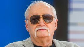 Mężczyzna w okularach przeciwsłonecznych z białymi wąsami, w szarym garniturze, na tle niebieskim.