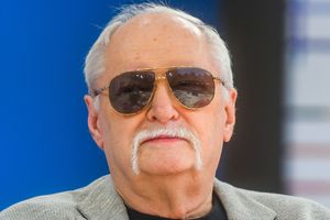 Mężczyzna w okularach przeciwsłonecznych z białymi wąsami, w szarym garniturze, na tle niebieskim.