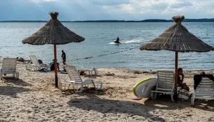 Plaża z parasolami, leżakami, ludźmi na piasku i skutrem wodnym na tle jeziora.