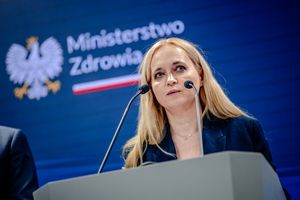 Na konferencji prasowej stoi kobieta w niebieskiej marynarce, mówi do mikrofonu.