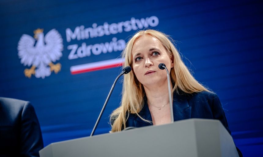 Na konferencji prasowej stoi kobieta w niebieskiej marynarce, mówi do mikrofonu.
