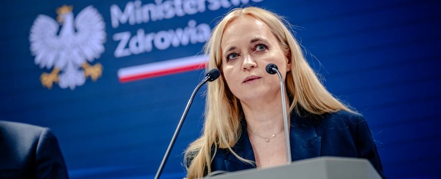 Na konferencji prasowej stoi kobieta w niebieskiej marynarce, mówi do mikrofonu.