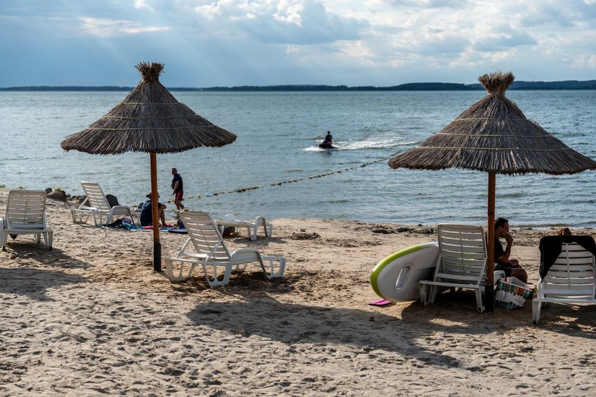 Plaża z parasolami, leżakami i w tle osoba na skuterze wodnym oraz spokojne wody jeziora.