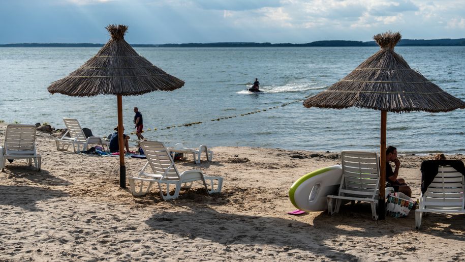 Plaża z parasolami, leżakami i w tle osoba na skuterze wodnym oraz spokojne wody jeziora.
