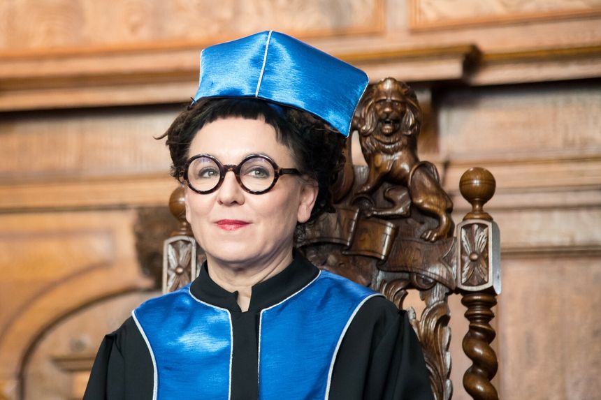 Kobieta w akademickiej todze i birecie, siedząca na ozdobnym tronie w historycznej sali.