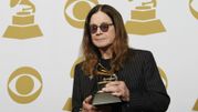 Mężczyzna w ciemnych okularach trzyma nagrodę Grammy na tle złotych symboli gramofonu.