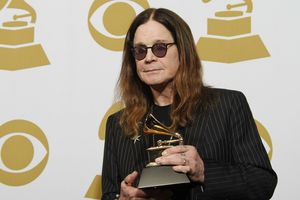 Mężczyzna w ciemnych okularach trzyma nagrodę Grammy na tle złotych symboli gramofonu.
