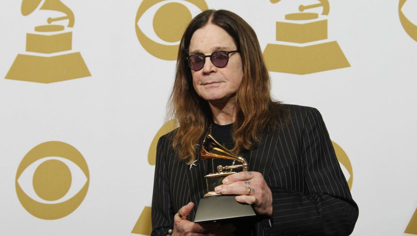 Mężczyzna w ciemnych okularach trzyma nagrodę Grammy na tle złotych symboli gramofonu.