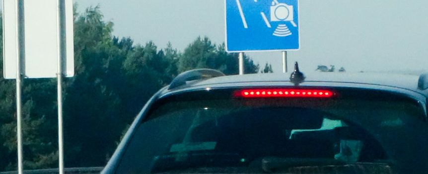 Zdjęcie przedstawia tył samochodu przed znakiem drogowym z kamerą monitorującą ruch.