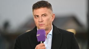 Mateusz Borek w czarnym płaszczu trzyma mikrofon i mówi, w tle rozmyte światła.