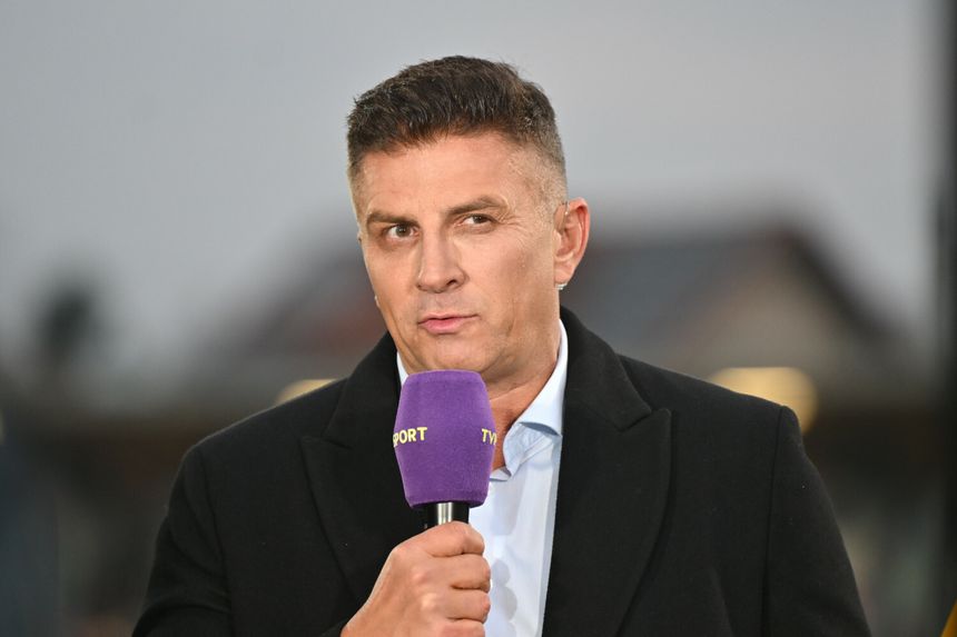 Mateusz Borek w czarnym płaszczu trzyma mikrofon i mówi, w tle rozmyte światła.