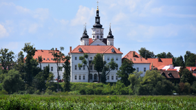 Piękny, biały pałac z czerwonymi dachami otoczony zielenią, w tle błękitne niebo.