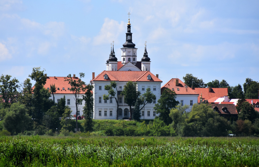 Piękny, biały pałac z czerwonymi dachami otoczony zielenią, w tle błękitne niebo.