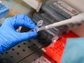 Ręka w rękawiczce trzyma pipetę, pobierając próbkę z laboratorium. W tle zdjęcia widoczne pojemniki.