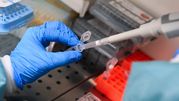 Ręka w rękawiczce trzyma pipetę, pobierając próbkę z laboratorium. W tle zdjęcia widoczne pojemniki.