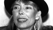 Joni Mitchell z szerokim uśmiechem, w kapeluszu, z dużymi kolczykami.