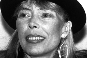 Joni Mitchell z szerokim uśmiechem, w kapeluszu, z dużymi kolczykami.