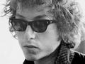Bob Dylan w ciemnych okularach, z kręconymi włosami, wyrazem skupienia na twarzy.