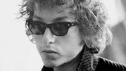 Bob Dylan w ciemnych okularach, z kręconymi włosami, wyrazem skupienia na twarzy.