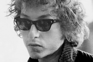 Bob Dylan w ciemnych okularach, z kręconymi włosami, wyrazem skupienia na twarzy.
