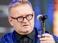 Mężczyzna w okularach trzyma mikrofon, w tle kolorowe światła. Niezidentyfikowany polityk.