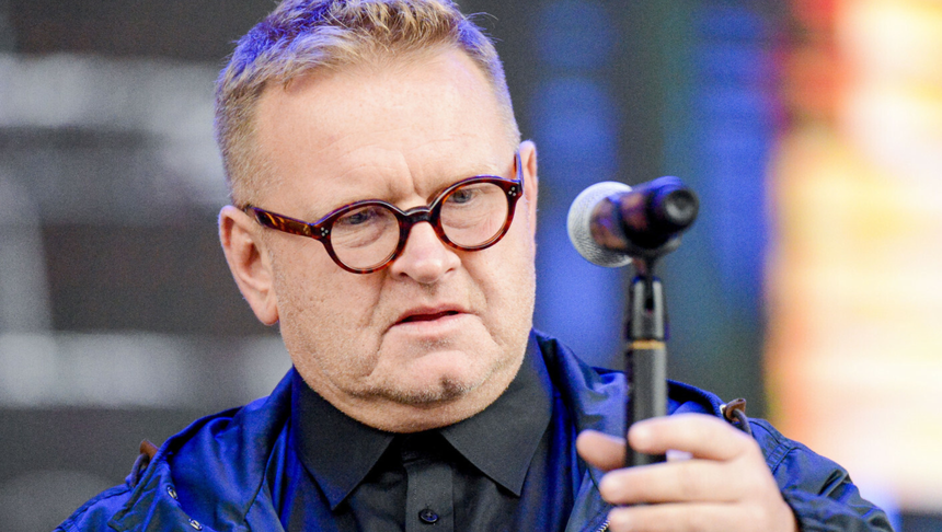 Mężczyzna w okularach trzyma mikrofon, w tle kolorowe światła. Niezidentyfikowany polityk.
