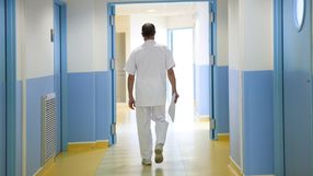 Person w białym uniformie medycznym idzie korytarzem szpitalnym z niebieskimi ścianami.
