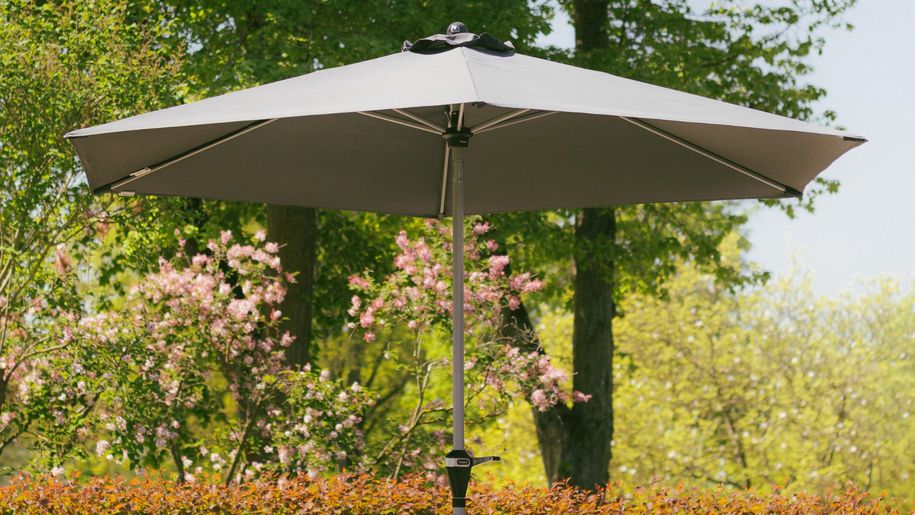 Na zdjęciu szary parasol ogrodowy w otoczeniu zieleni i kwitnących drzew.