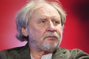 Na zdjęciu mężczyzna z siwymi włosami i brodą, w ciemnym swetrze, na tle w kolorze czerwonym.