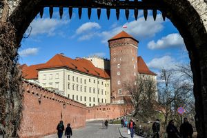 Widok na zamek Wawelski przez bramę oraz spacerujących ludzi w słoneczny dzień.
