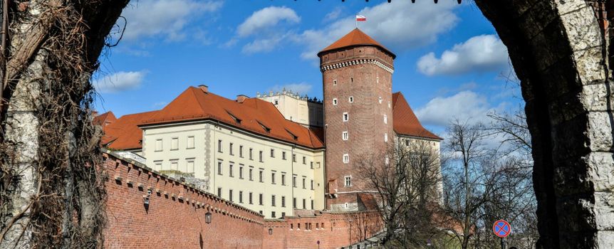 Widok na zamek Wawelski przez bramę oraz spacerujących ludzi w słoneczny dzień.
