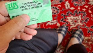 Na zdjęciu widoczna jest ręka trzymająca zieloną legitymację emeryta-rencisty, w tle dywan.