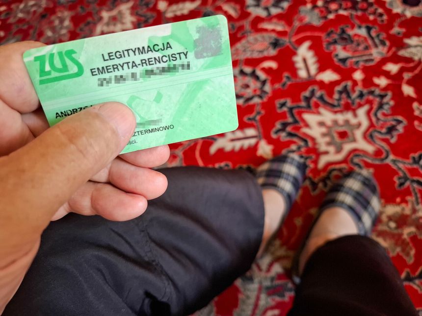 Na zdjęciu widoczna jest ręka trzymająca zieloną legitymację emeryta-rencisty, w tle dywan.