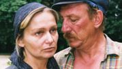Na zdjęciu para, mężczyzna z wąsem blisko kobiety w chustce, w tle zieleń drzew.