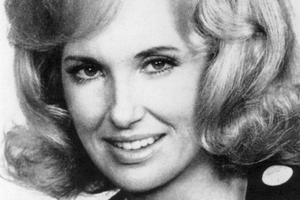 Na zdjęciu uśmiechnięta Tammy Wynette o falowanych, jasnych włosach i czarnej bluzce w białe kropki.