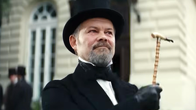 Mężczyzna w cylinderze i fraku trzyma laskę, uśmiechnięty, na tle budynku.