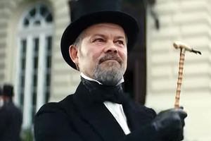 Mężczyzna w cylinderze i fraku trzyma laskę, uśmiechnięty, na tle budynku.