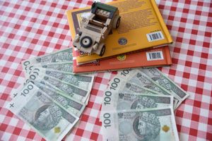 Na czerwono-białym tle leży gotówka, samochód z klocków oraz książki.
