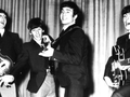 Muzycy z zespołu The Beatles w garniturach grają na instrumentach, śpiewając na scenie.