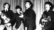 Muzycy z zespołu The Beatles w garniturach grają na instrumentach, śpiewając na scenie.