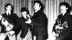 Muzycy z zespołu The Beatles w garniturach grają na instrumentach, śpiewając na scenie.