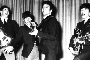 Muzycy z zespołu The Beatles w garniturach grają na instrumentach, śpiewając na scenie.