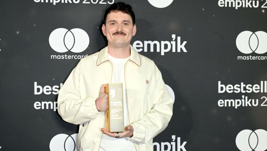 Mężczyzna w jasnej kurtce trzyma nagrodę na tle ciemnej ścianki z logo Empiku.